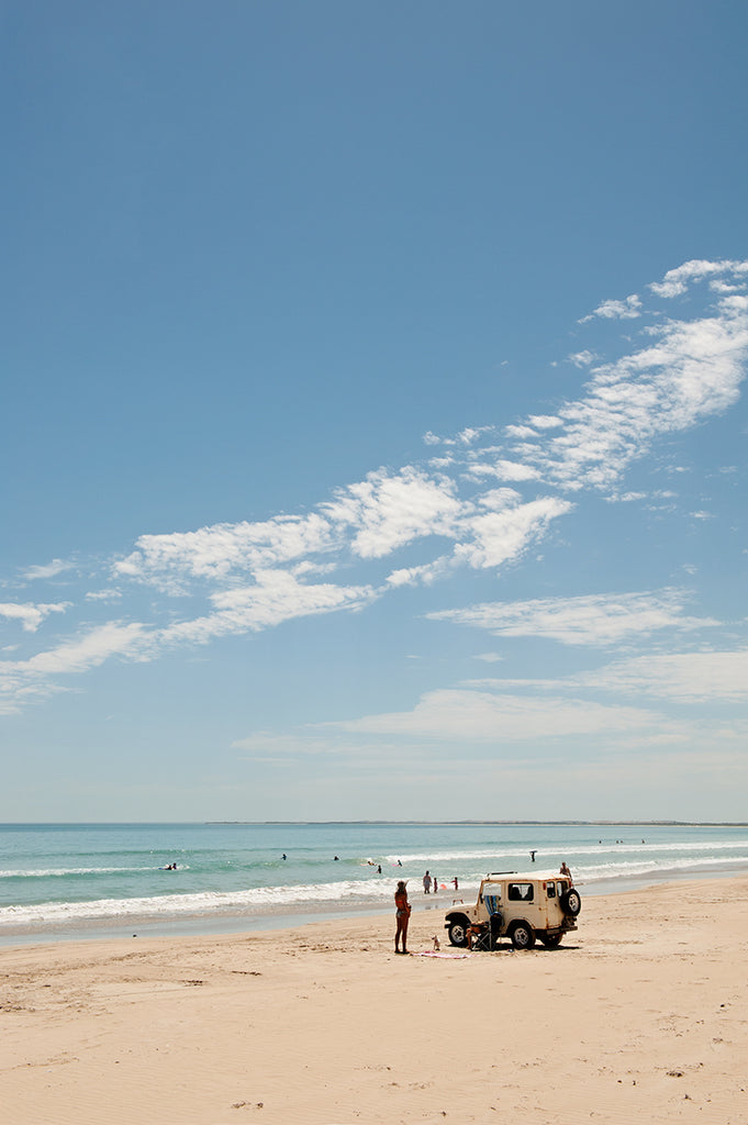 Australian beach print featuring a hot summer day on the beach with a land rover parked on the sand and a girl and her dog, swimmers in the blue ocean with a blue sky overhead