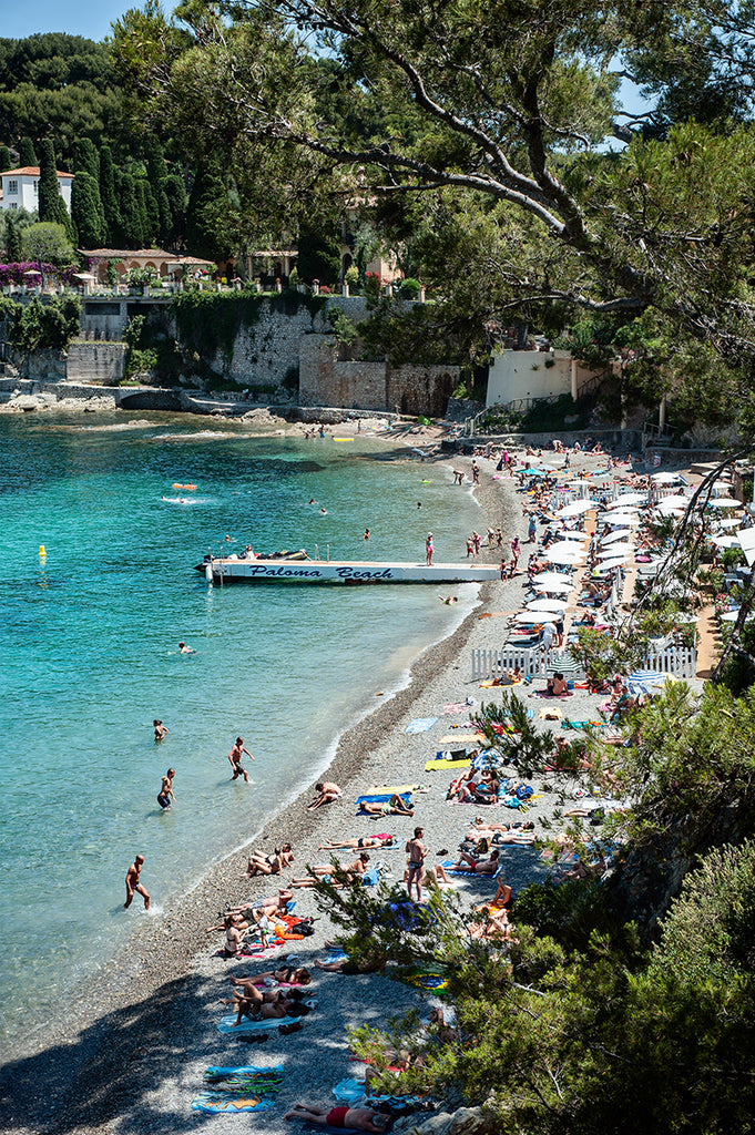 Paloma Beach is a French Riviera fine art limited edition print featuring the small swimming cove with its beachgoers in the water, the Paloma Beach jetty and the private beach with its sun umbrellas and beachgoers Beach shot from above  on a busy hot summer day 