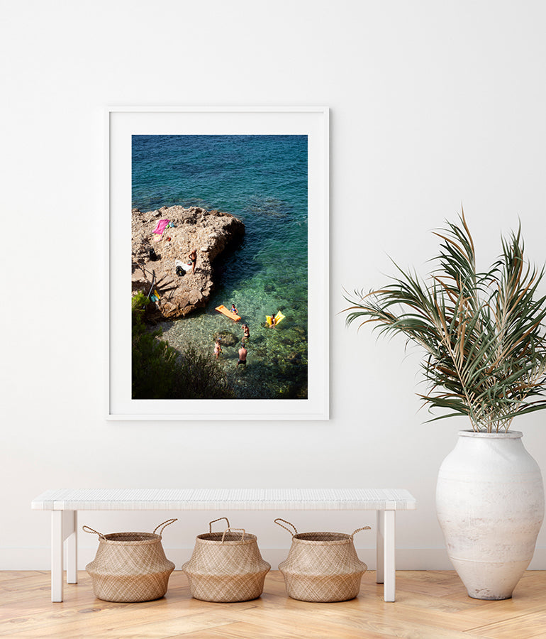 French Riviera print featuring people  in a beautiful swimming cove shot from above showing people swimming and standing with their colourful air mattresses on the water as well as on the rocks. Part of the Into The Blue fine art print series by Millie Brown