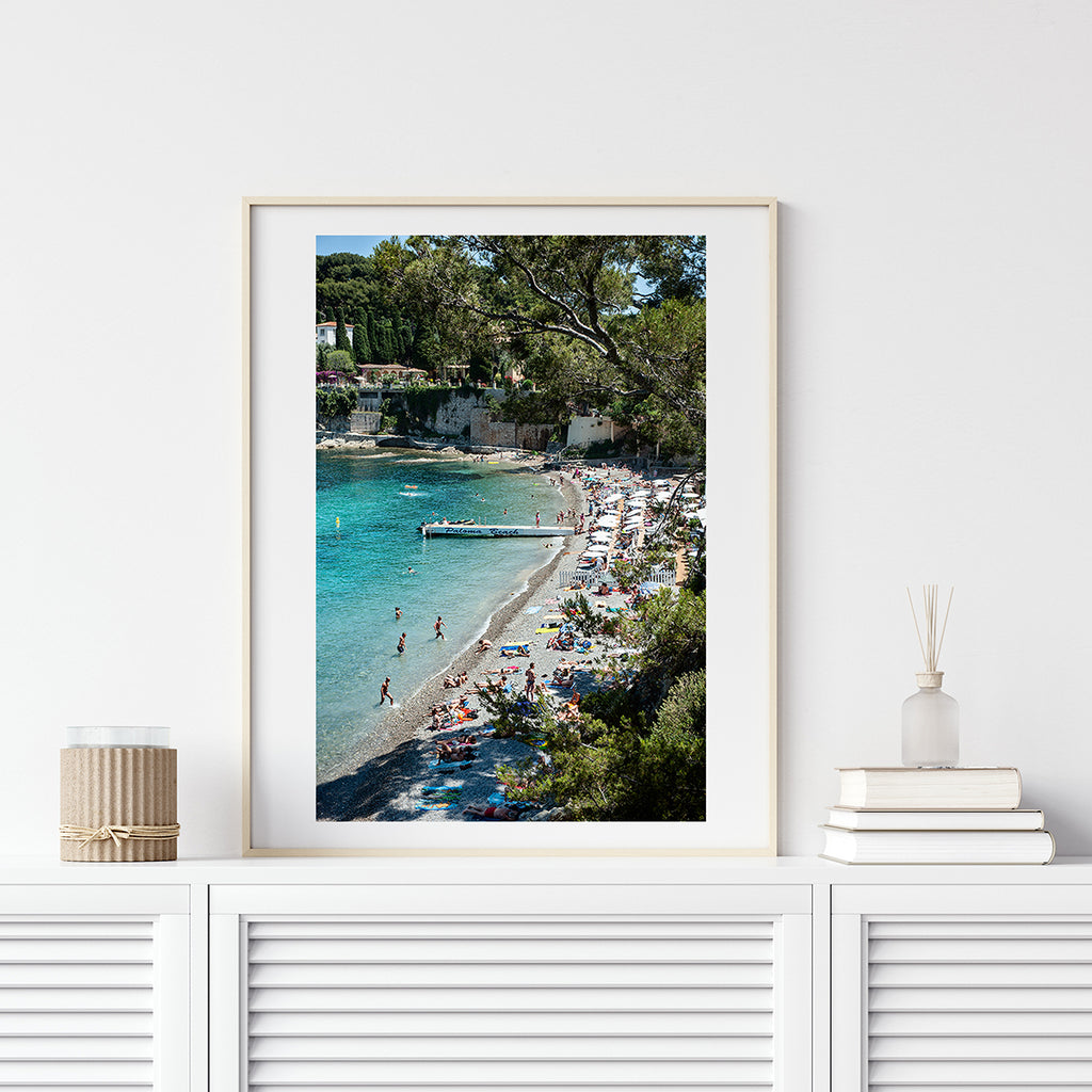 French Riviera beach print of Paloma Beach in summer. Photographed from above and featuring people in the blue water and on the beach. The swimming cove features the white Paloma Beach jetty and is surrounded by green vegetation