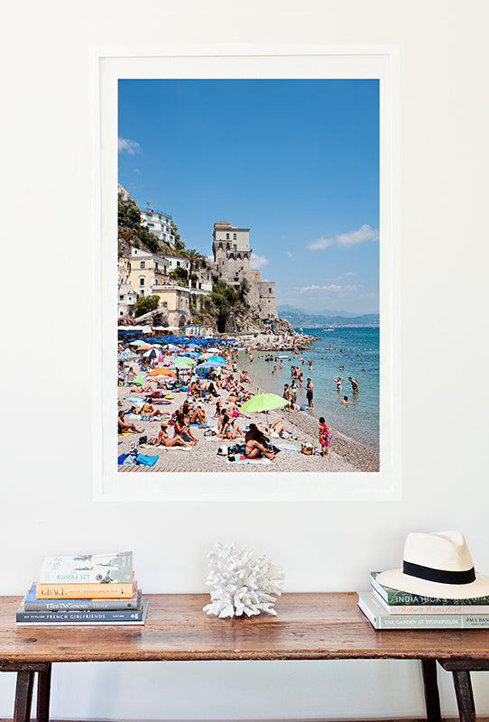 Amalfi coast photographic print of Cetara beach on the Amalfi Coast with colourful beach umbrellas and beachgoers on the shore and in the sea and an old stone building in the background by photographer Millie Brown
