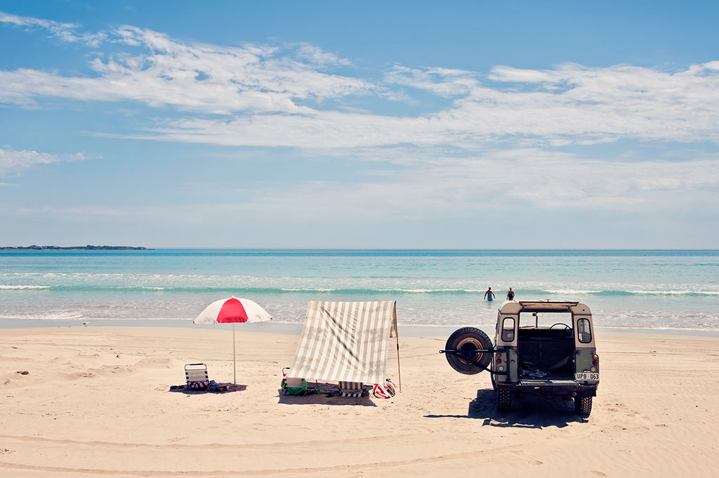 Australian Beach photographic print featuring beautiful long beach in Robe South Australia. The Land Rover s parked, the beach tent is pitched and the umbrella is up. The clear blue ocean entices a swimmer into the water on a hot summer day in Australia. Available as a fine art limited edition print by Millie Brown