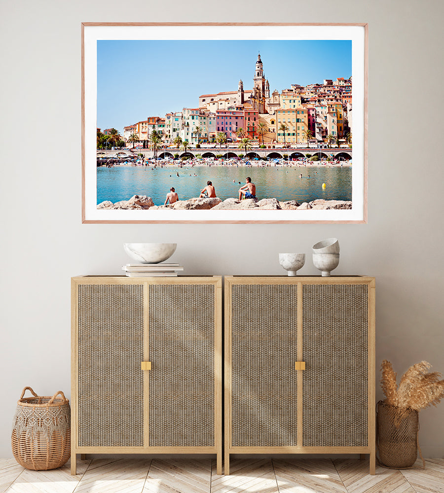 French Riviera print featuring the beautiful colourful village of Menton and it's beach with the blue mediterranean in the foreground and swimmers on the rocks by professional photographer Millie Brown