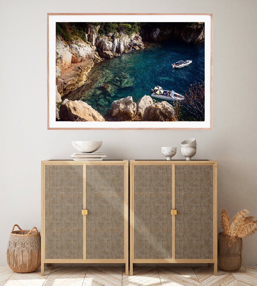 French Riviera Fine Art Wall print of the blue mediterranean sea in a small cove on Cap Ferrat with a person sitting on the rocks and two leisure boats with people sun bathing by travel photographer Millie Brown