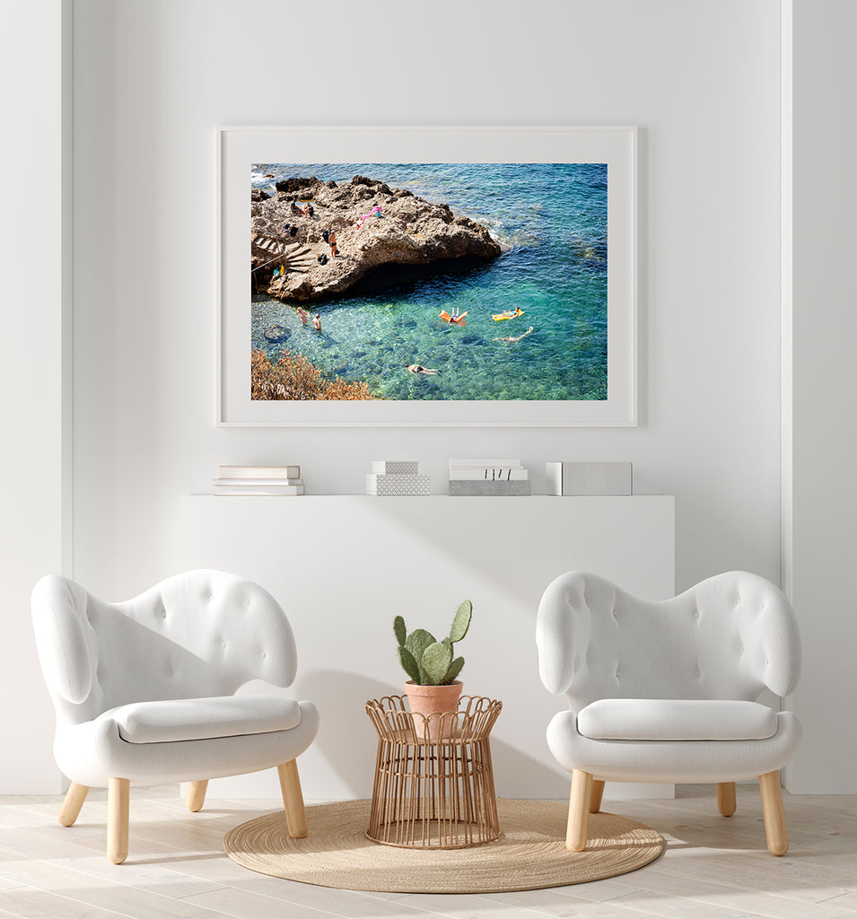 French Riviera beach print featuring people swimming in a cove of beautiful blue water with a rocky outcrop and rocky stairs leading down to the sea with a swimmer standing on it looking down at the swimmers