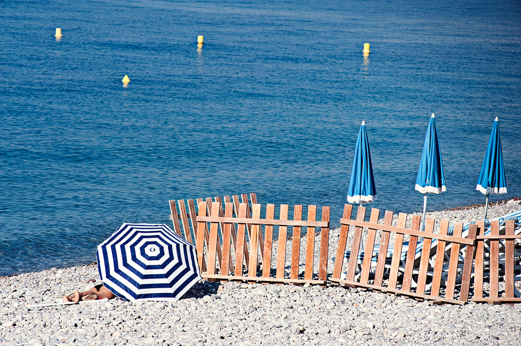 French Riviera Print featuring the pebbly beach in Nice France and a blue and blue beach umbrellas next to the blue mediterranean sea