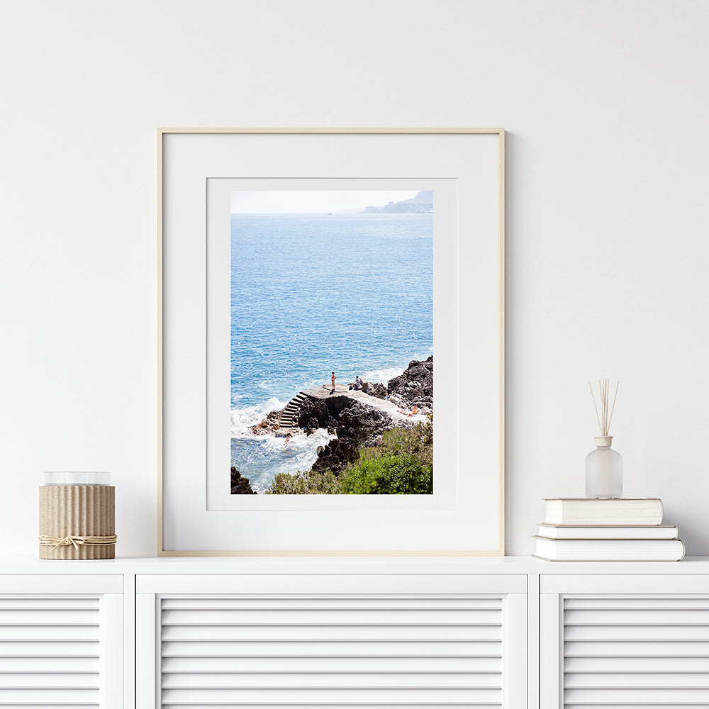 French Riviera beach wall art. A girl in a red bikini stands down below on the rocks looking out to the Mediterranean Sea on the French Riviera, rocky steps lead down to the water by Photographer Millie Brown