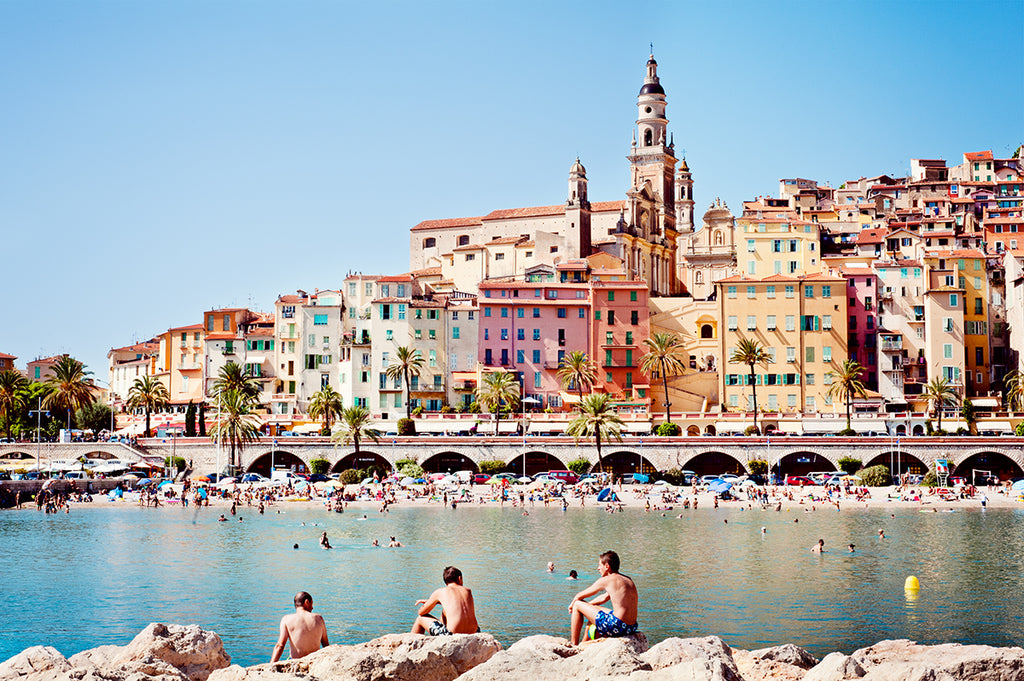 French Riviera Fine Art Print featuring the beautiful colourful village of Menton and it's beach with the blue mediterranean in the foreground and swimmers on the rocks by professional photographer Millie Brown