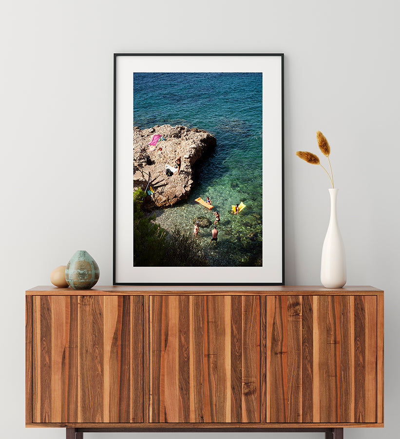 Beach Photography print French Riviera swimming cove shot from above showing people swimming and standing with their colourful air mattresses on the water as well as on the rocks. Part of the Into The Blue fine art print series by Millie Brown