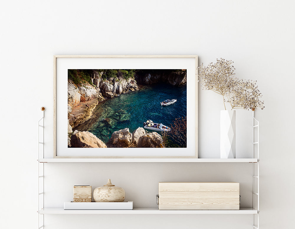 French Riviera summer print featuring a small cove on Cap Ferrat peninsula with two boats on the water and one person sitting on the rocky shoreline on a hot summer day. Shot from above.