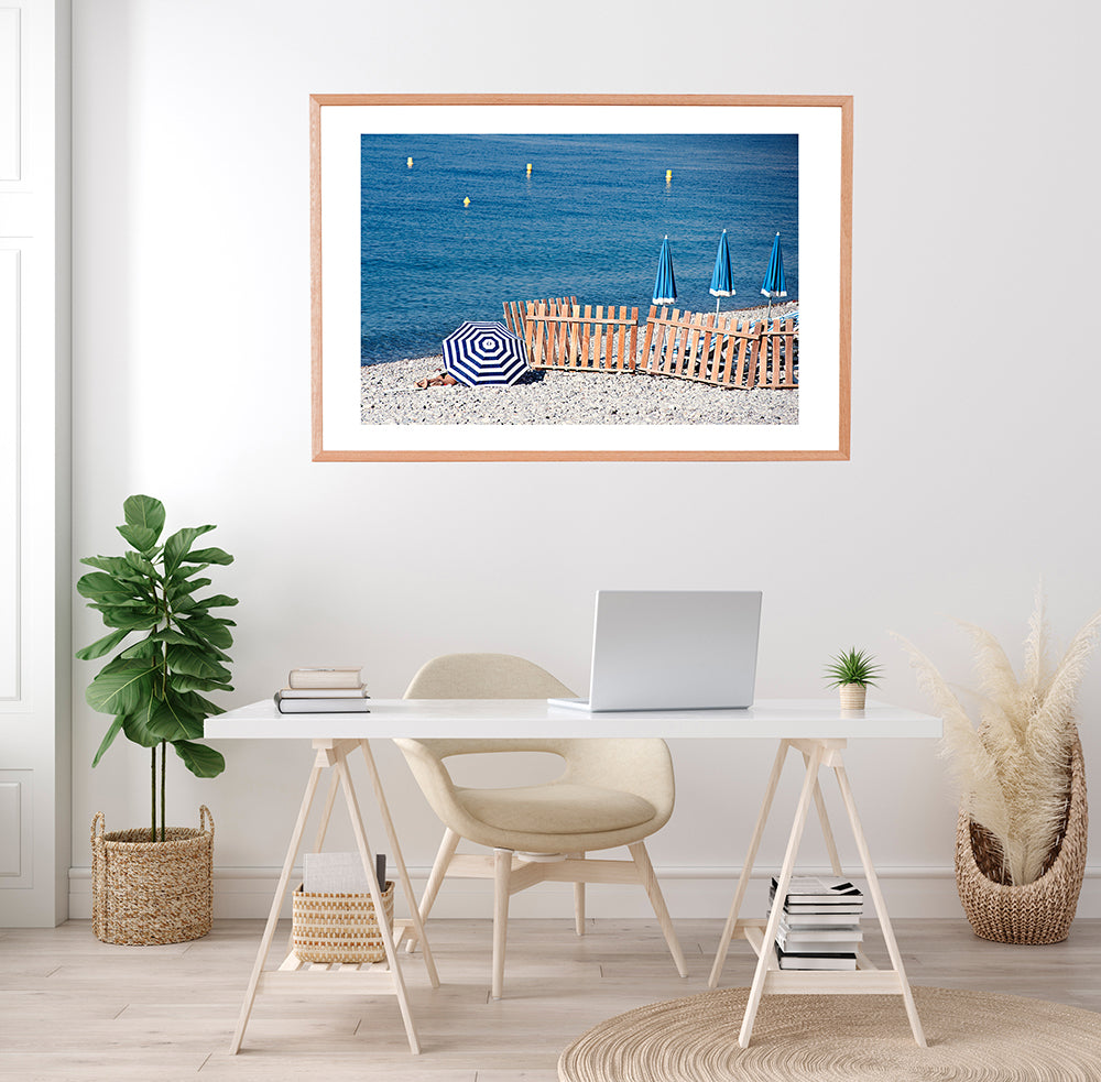 French Riviera Wall Art Print featuring the pebbly beach in Nice France and a blue and blue beach umbrellas next to the blue mediterranean sea