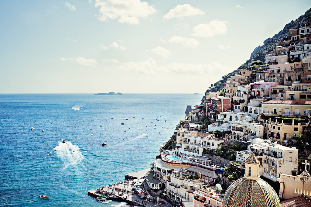 Amalfi Coast print featuring Positano village hanging off a cliff falling into the blue mediterranean sea and showing the small boats moving along the water by Millie Brown