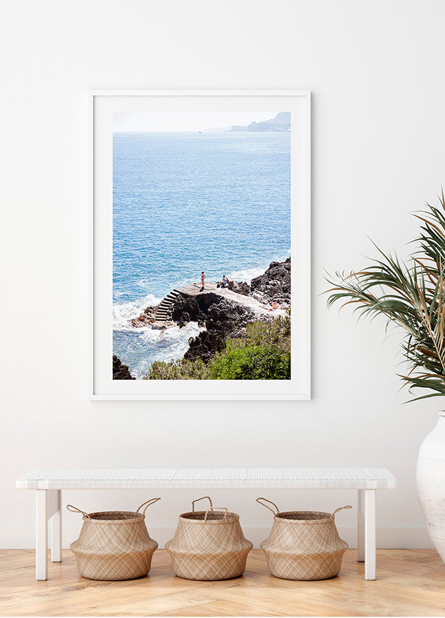 French Riviera beach wall art. A girl in a red bikini stands down below on the rocks looking out to the Mediterranean Sea on the French Riviera, rocky steps lead down to the water  by Photographer Millie Brown
